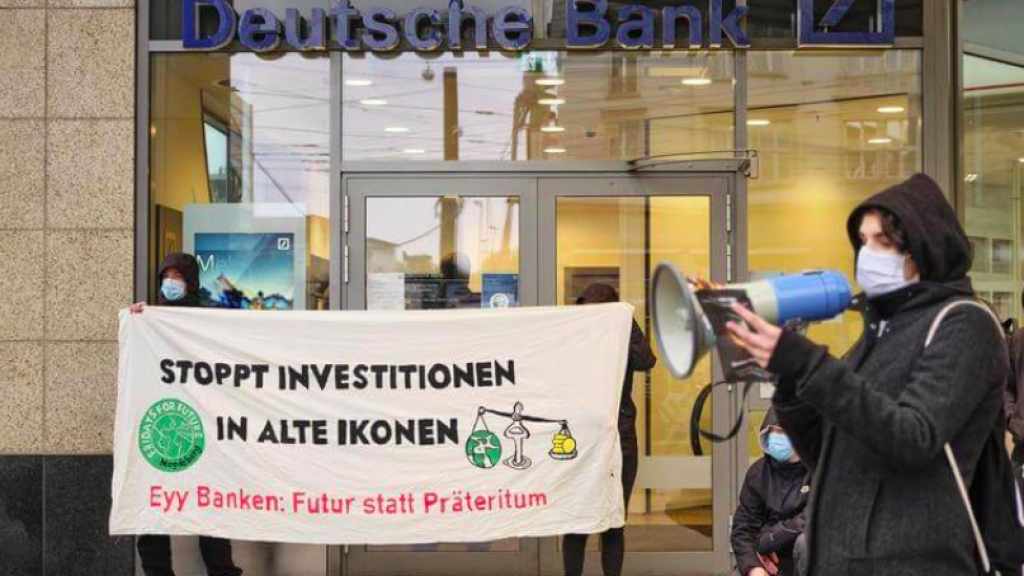 Menschen protestieren vor einer Deutschen Bank Filiale mit einem Banner auf dem steht "Stoppt Investitionen in alte Ikonen"